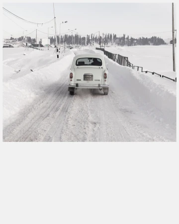 Rear view of a white vintage car driving on a snow-covered road with trees in the background during winter under a clear sky.