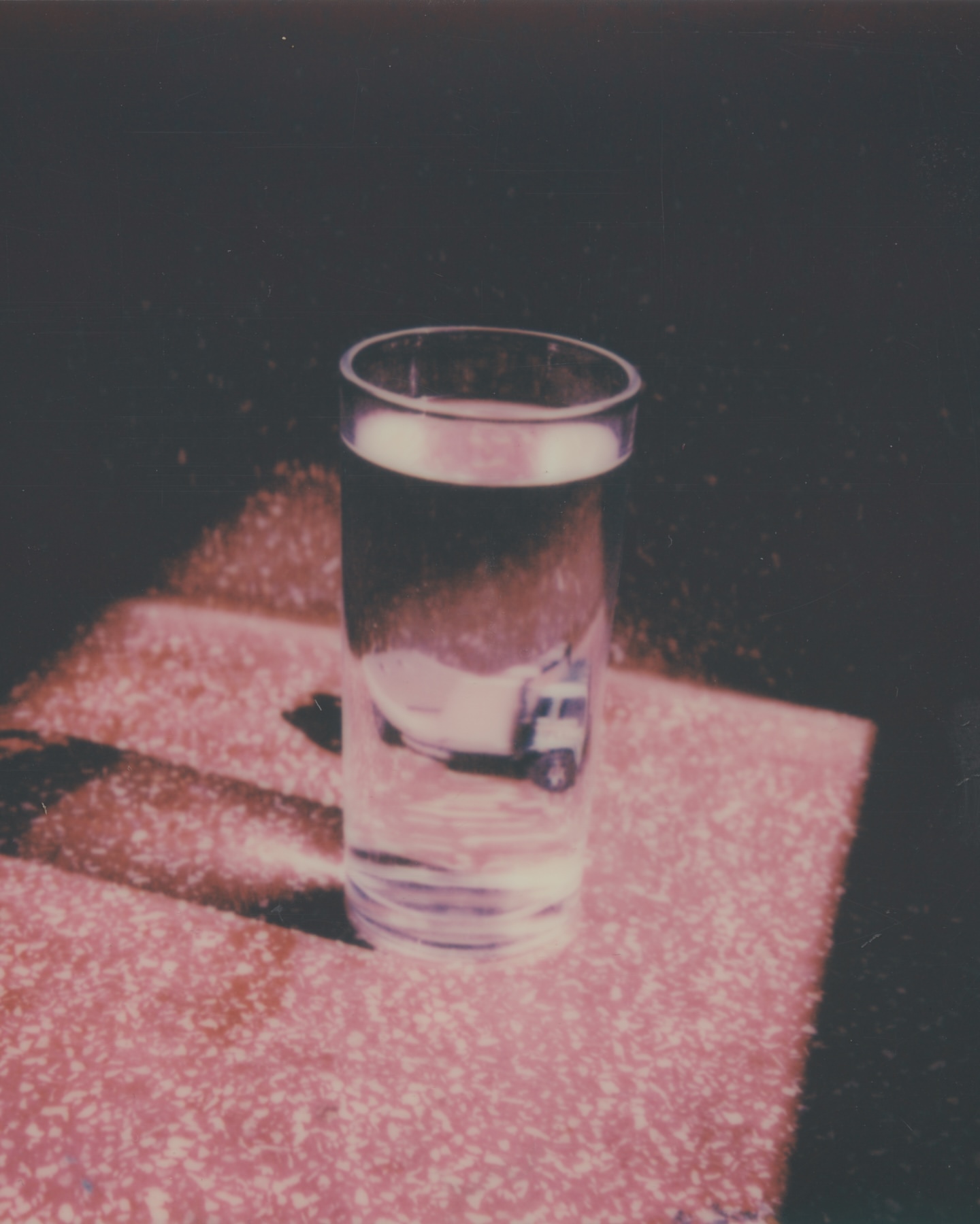 Close-up studio shot of a clear glass filled with water, casting shadows on a speckled peach-colored surface against a dark background.