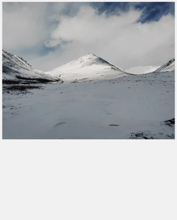 Snow-covered mountain range under a cloudy sky, showcasing a stark and serene winter landscape with muted tones.