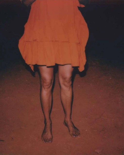 Woman in an orange dress stands barefoot on dark ground. The low angle and dark background create a striking contrast and candid feel.