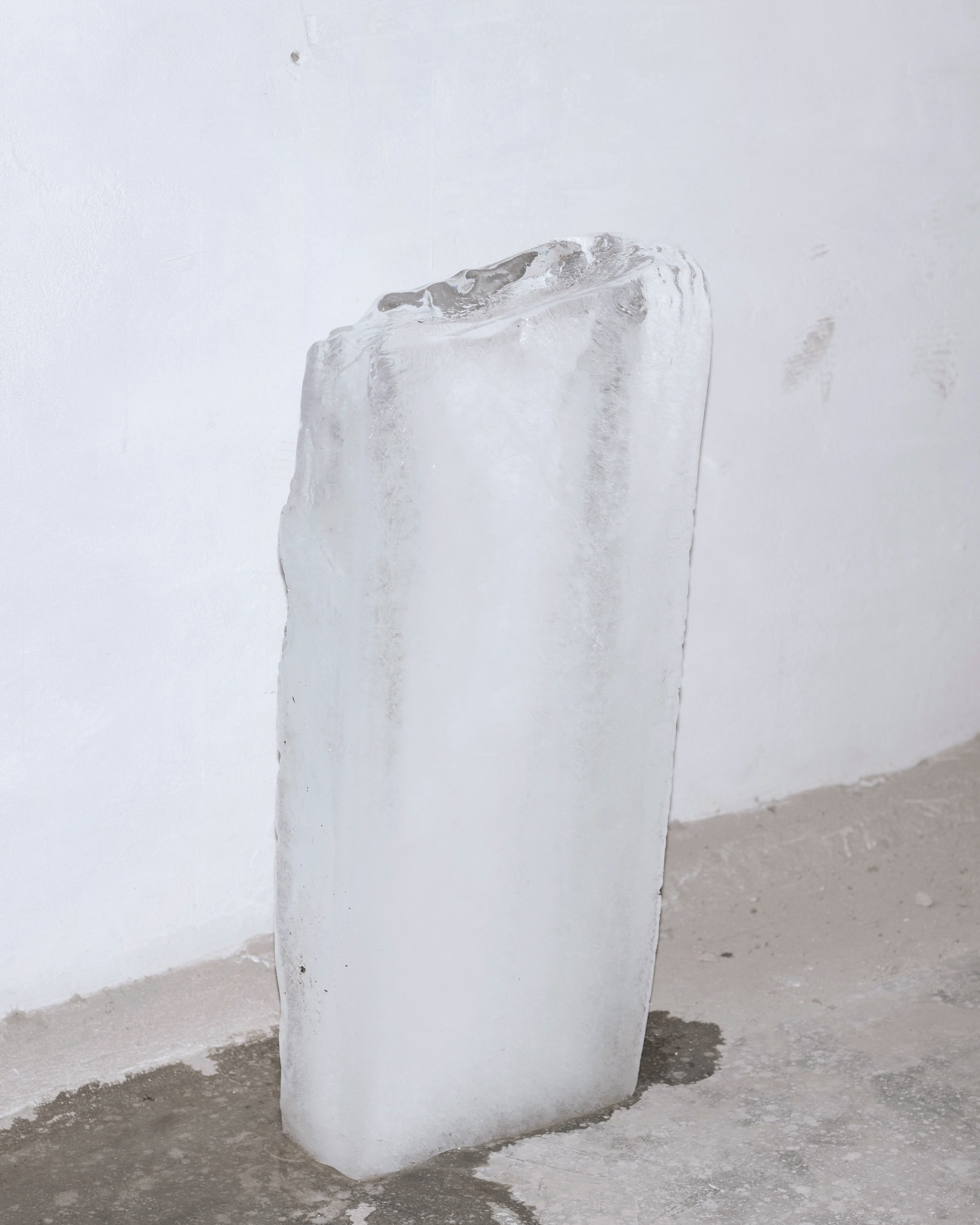 Icy block with melting streaks stands indoors against white wall, reflecting light on concrete floor with puddles in austere scene.