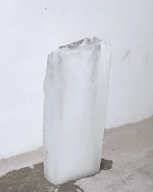 Icy block with melting streaks stands indoors against white wall, reflecting light on concrete floor with puddles in austere scene.