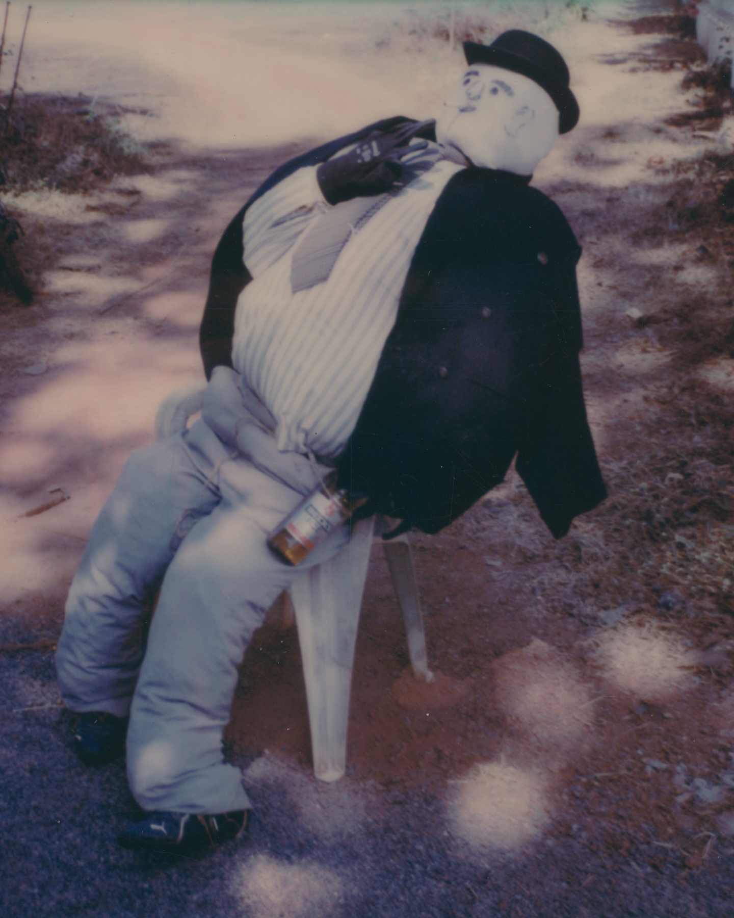 Mannequin seated on a chair outdoors wearing a suit and hat, holding a bottle, in a vintage-style photograph with faded colors.