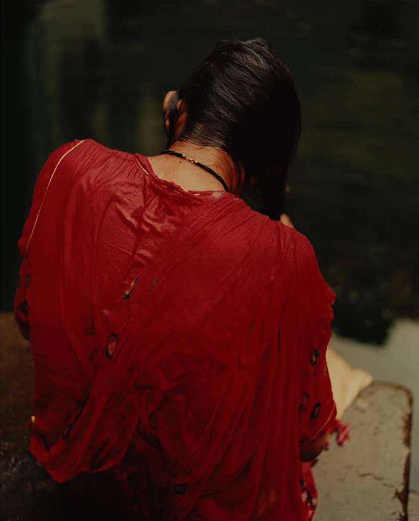 Back view of an adult woman in a red dress at an outdoor waterside setting, near a dark pool or lake, photographed in medium shot.