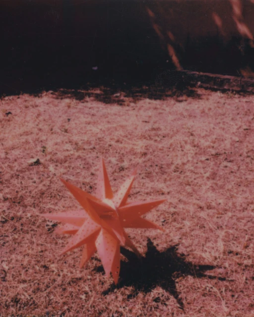 Abstract geometric star sculpture on grass in a minimalist, outdoor setting with muted colors. The object casts a shadow.