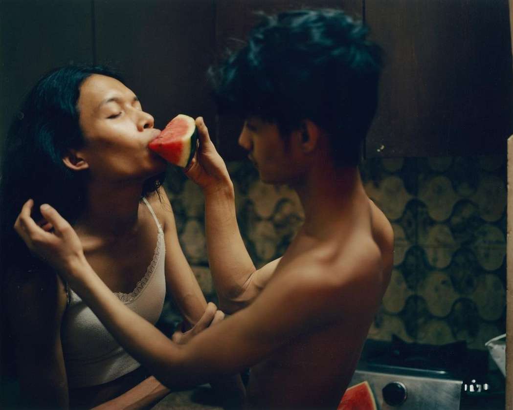 Woman eating watermelon slice held by a man, close up in an indoor setting with dark walls, soft lighting, and visible cooking range.