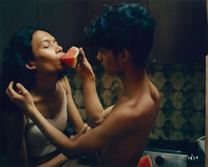 Woman eating watermelon slice held by a man, close up in an indoor setting with dark walls, soft lighting, and visible cooking range.
