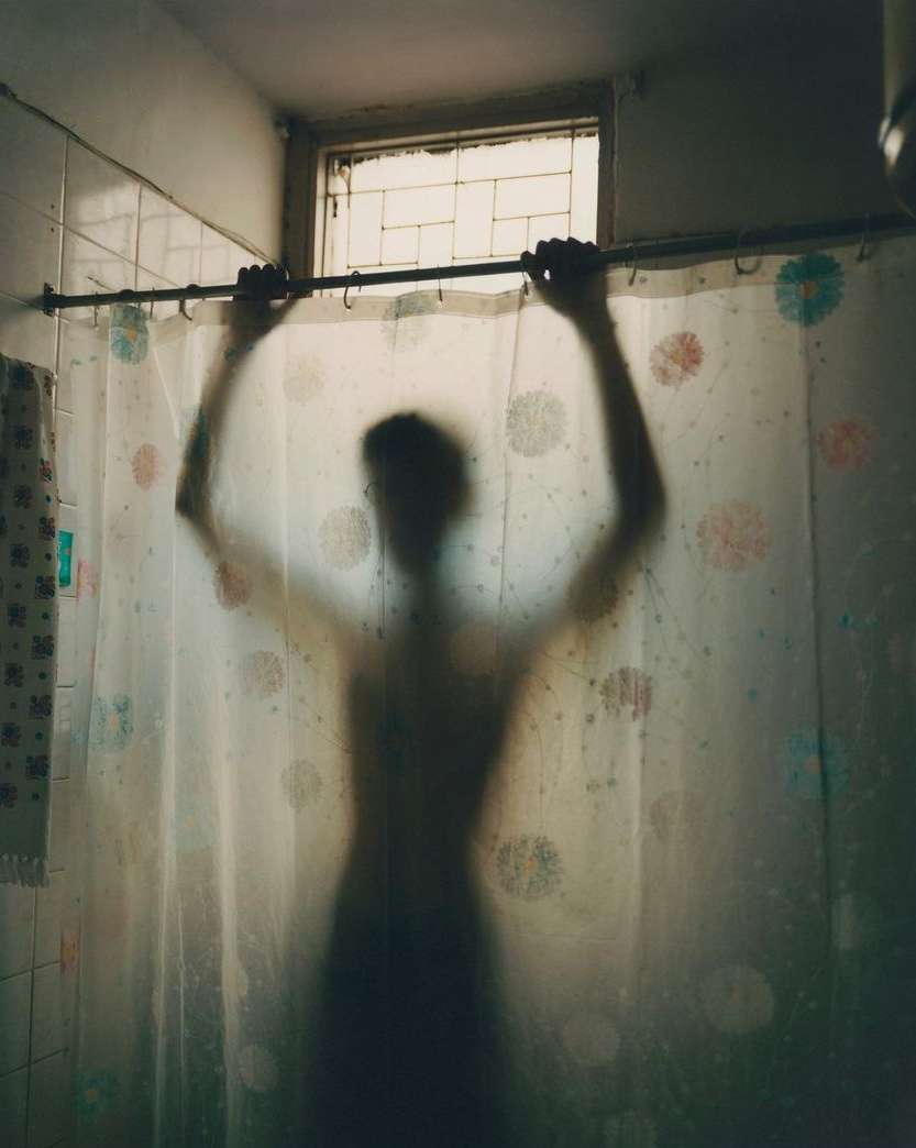 Silhouette of a figure behind a patterned shower curtain with hands on the shower curtain rod in a brightly lit bathroom.