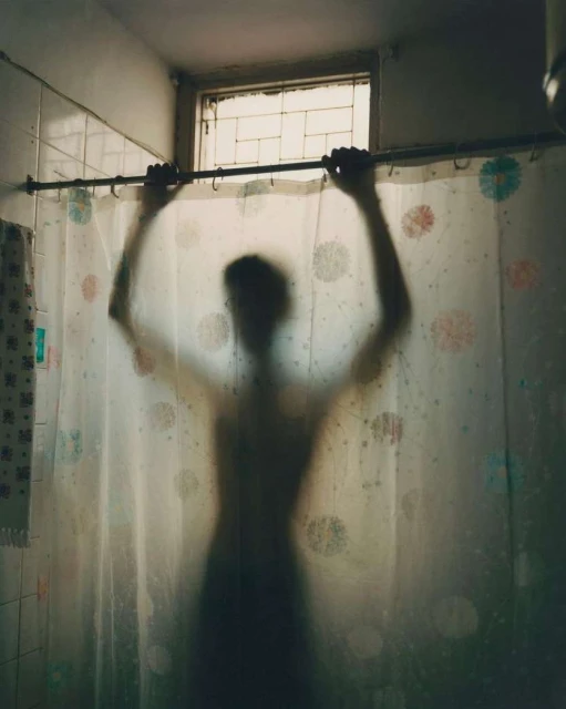 Silhouette of a figure behind a patterned shower curtain with hands on the shower curtain rod in a brightly lit bathroom.