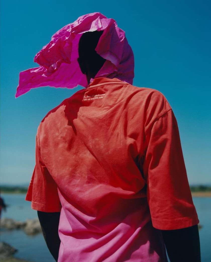 A person stands in a vibrant outdoor setting, wearing a red shirt and a pink head covering against a backdrop of clear blue sky and water.