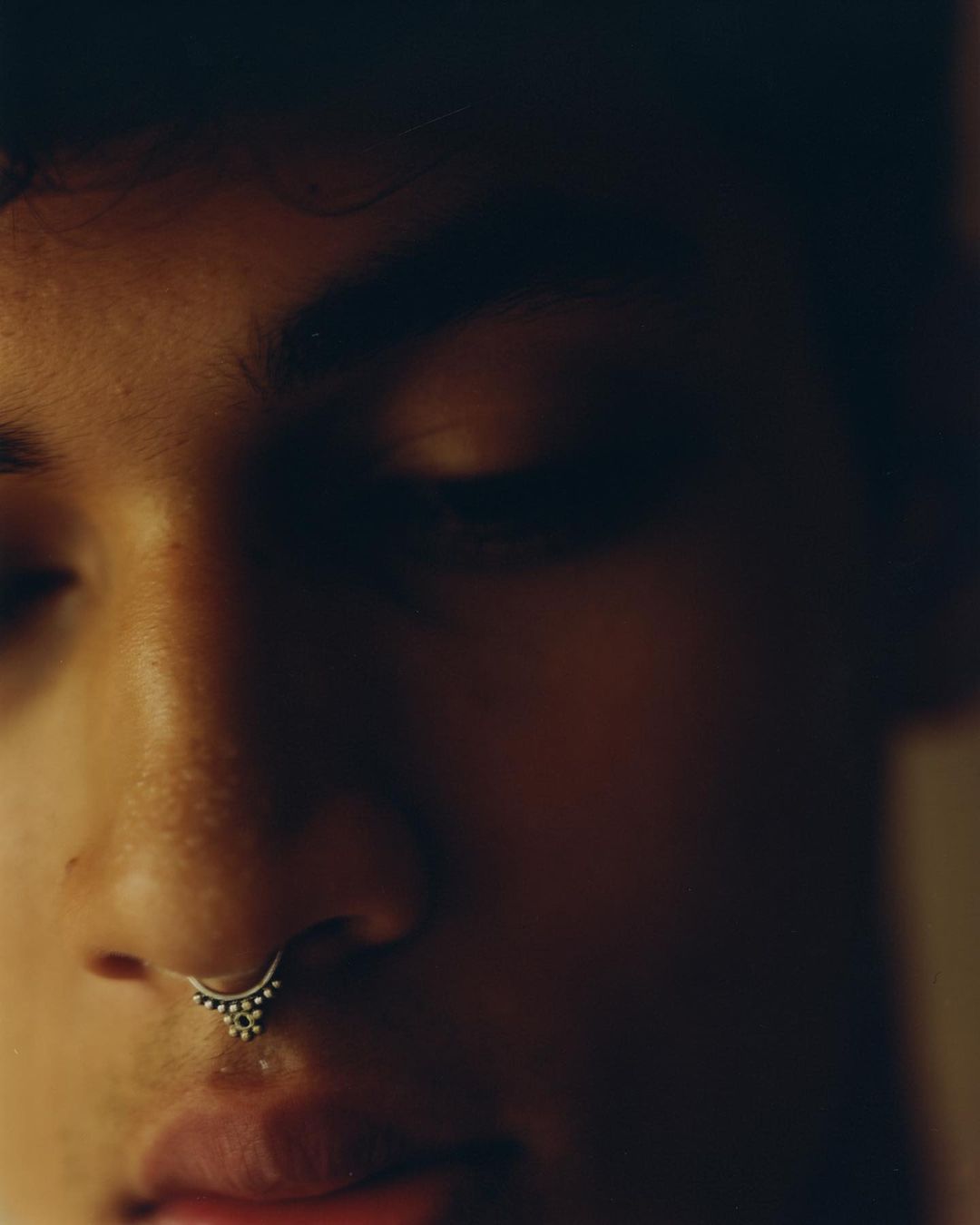 Close-up studio portrait of a young adult male with dark hair and a nose ring, in warm lighting and soft focus.