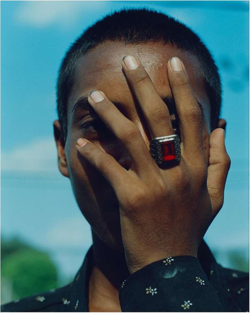 Close-up portrait of a Black adult man with short hair partially covering his face with his hand, adorned with a ruby ring, against a blue sky.