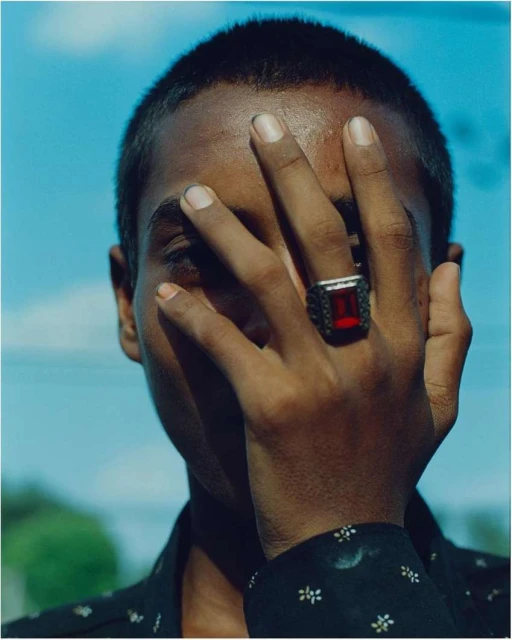 Close-up portrait of a Black adult man with short hair partially covering his face with his hand, adorned with a ruby ring, against a blue sky.