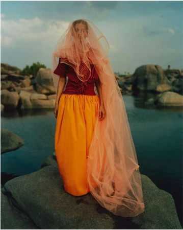 Model standing on a rock by water wearing a red striped top, orange skirt, and a sheer peach veil over head.