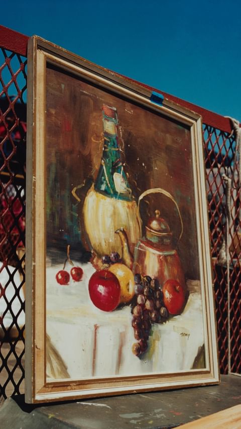 Still life painting of a wine bottle, kettle, and fruits on a table with a white cloth, set against a bright blue sky.