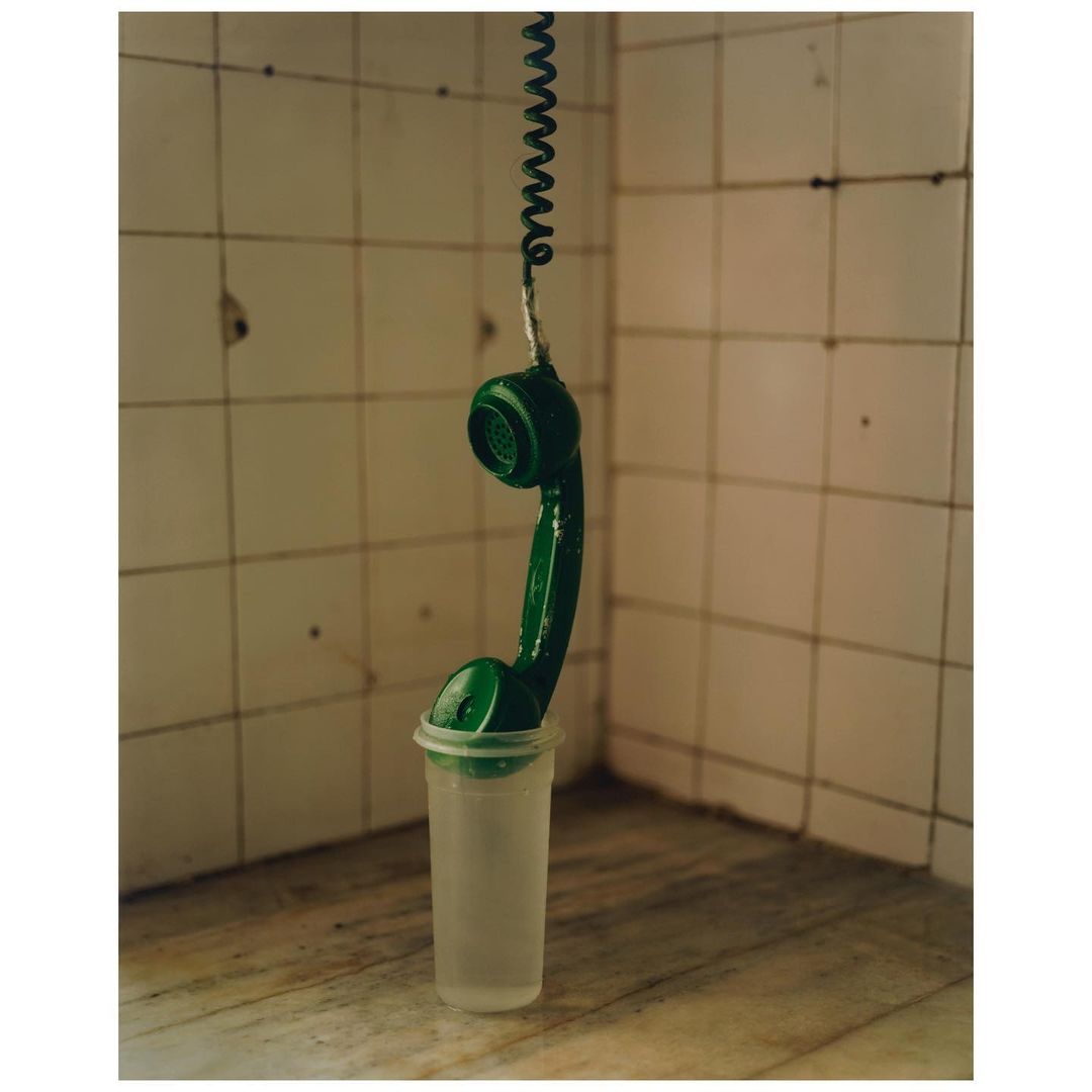 A green telephone receiver hanging from a coiled cord, suspended in a translucent plastic cup, set against a tiled wall and marble surface.
