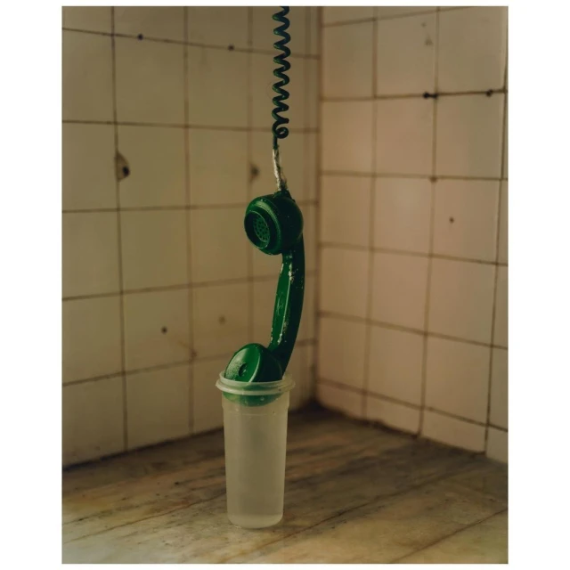A green telephone receiver hanging from a coiled cord, suspended in a translucent plastic cup, set against a tiled wall and marble surface.