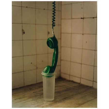 A green telephone receiver hanging from a coiled cord, suspended in a translucent plastic cup, set against a tiled wall and marble surface.