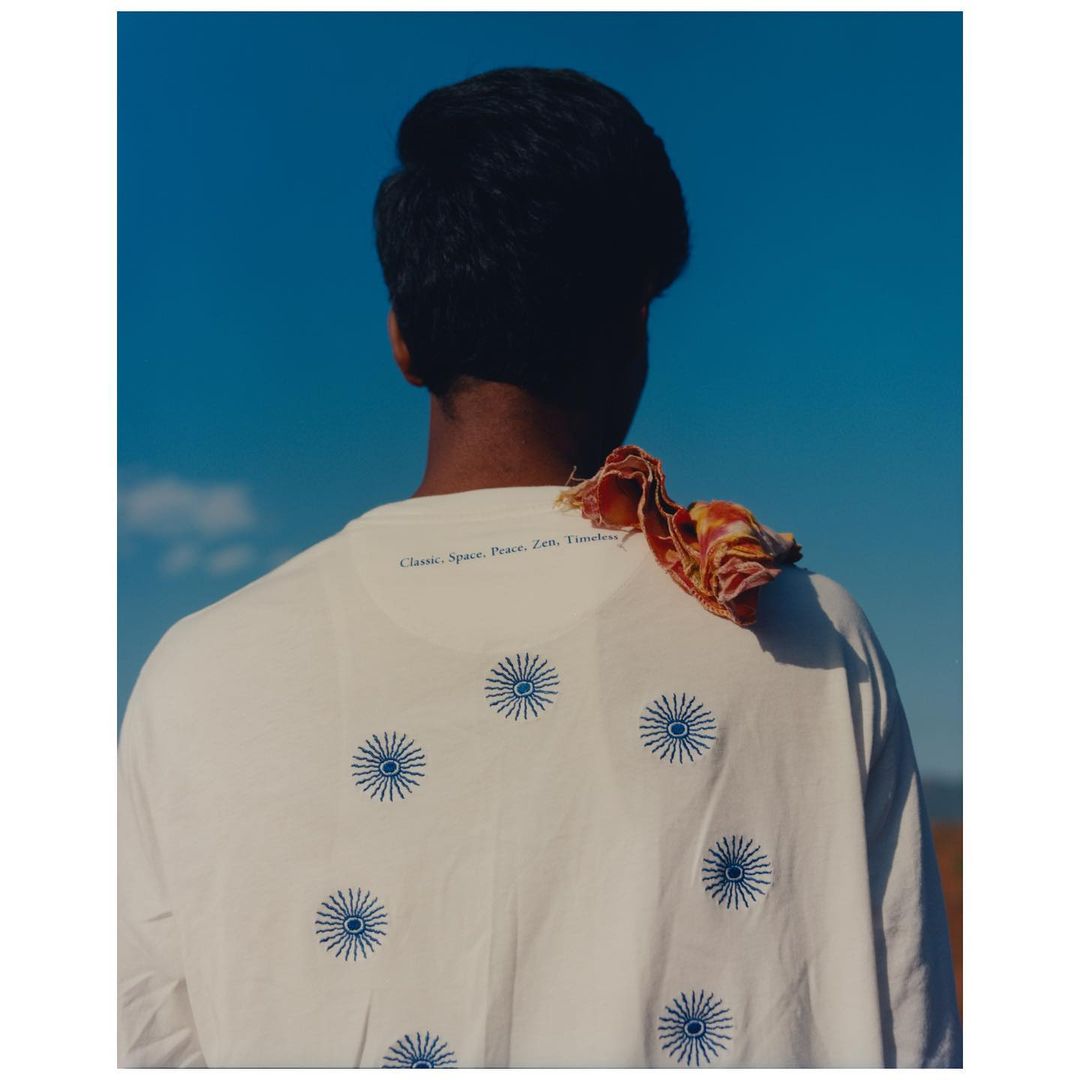 Back view of a person wearing a white t-shirt with blue sunbursts on it, against a clear blue sky, with a scarf draped on the shoulder.