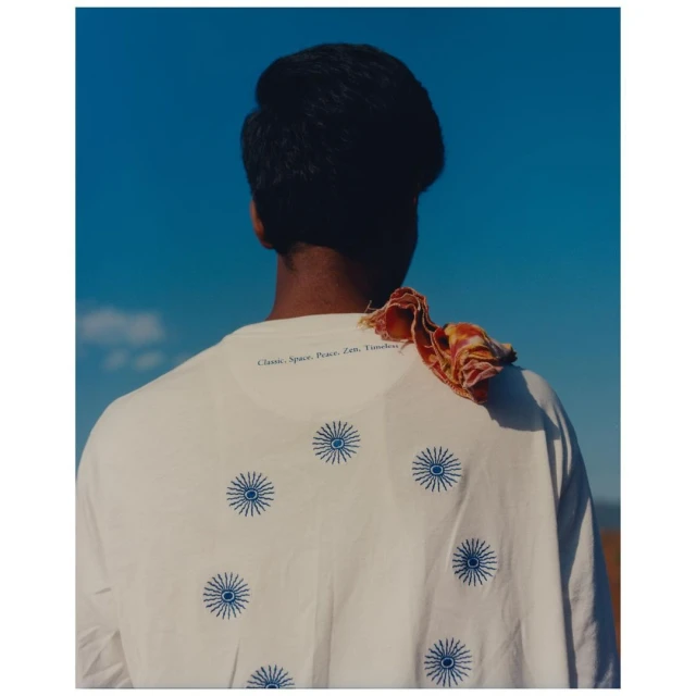Back view of a person wearing a white t-shirt with blue sunbursts on it, against a clear blue sky, with a scarf draped on the shoulder.