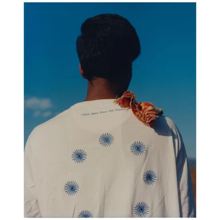 Back view of a person wearing a white t-shirt with blue sunbursts on it, against a clear blue sky, with a scarf draped on the shoulder.