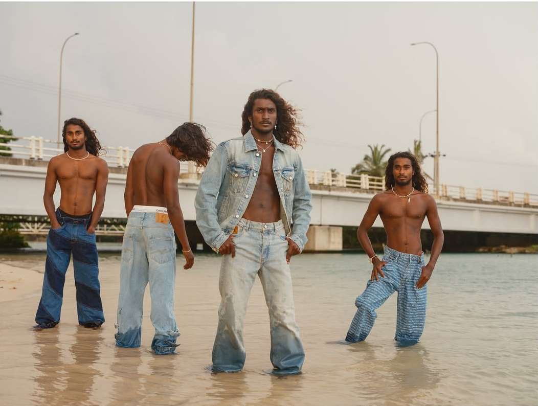 Four men with long dark hair standing in shallow water on a sandy beach wearing denim pants, photographed by Bharat Sikka.