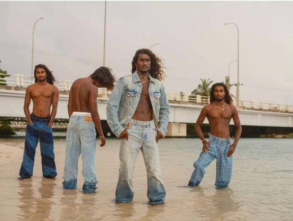 Four men with long dark hair standing in shallow water on a sandy beach wearing denim pants, photographed by Bharat Sikka.