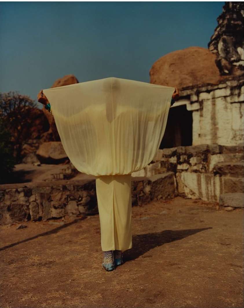 Person in yellow outfit holding a large, sheer fabric against a backdrop of ancient stone buildings and rocky terrain on a sunny day.