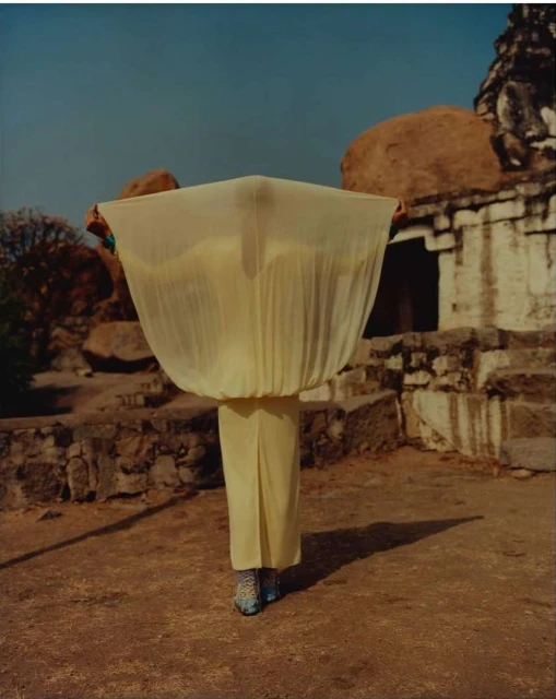 Person in yellow outfit holding a large, sheer fabric against a backdrop of ancient stone buildings and rocky terrain on a sunny day.