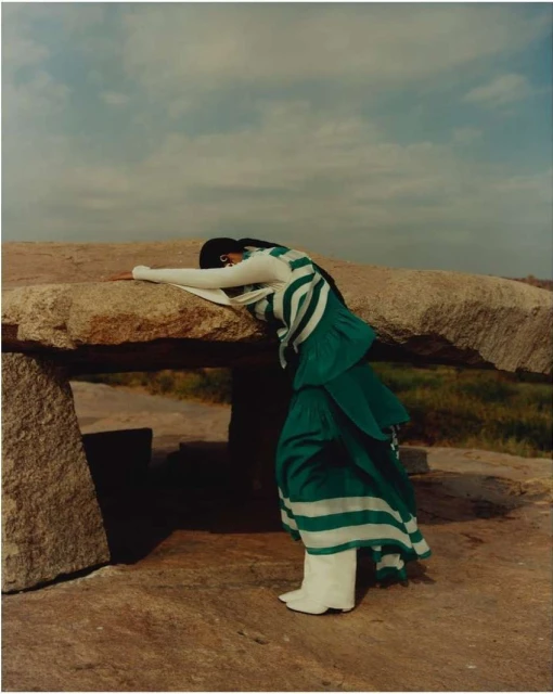 A woman in a green and white striped outfit leans on a stone structure in a rural outdoor scene under a cloudy sky.