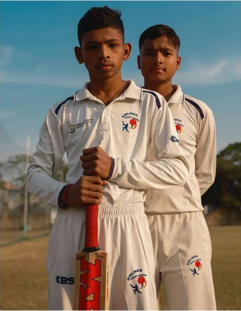 Two young cricket players wearing white uniforms hold a cricket bat outdoors with a clear blue sky in the background in a medium shot.