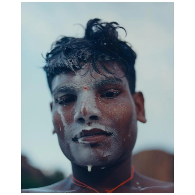 Close-up portrait of an Indian adult male with smeared white face paint and a red mark on his forehead, wearing an orange string necklace.