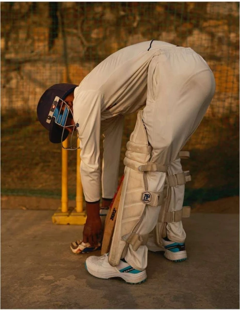 Cricket player in whites laces their shoe near wickets. The sportsman wears a helmet and pads. Gear and uniform dominate the indoor shot.