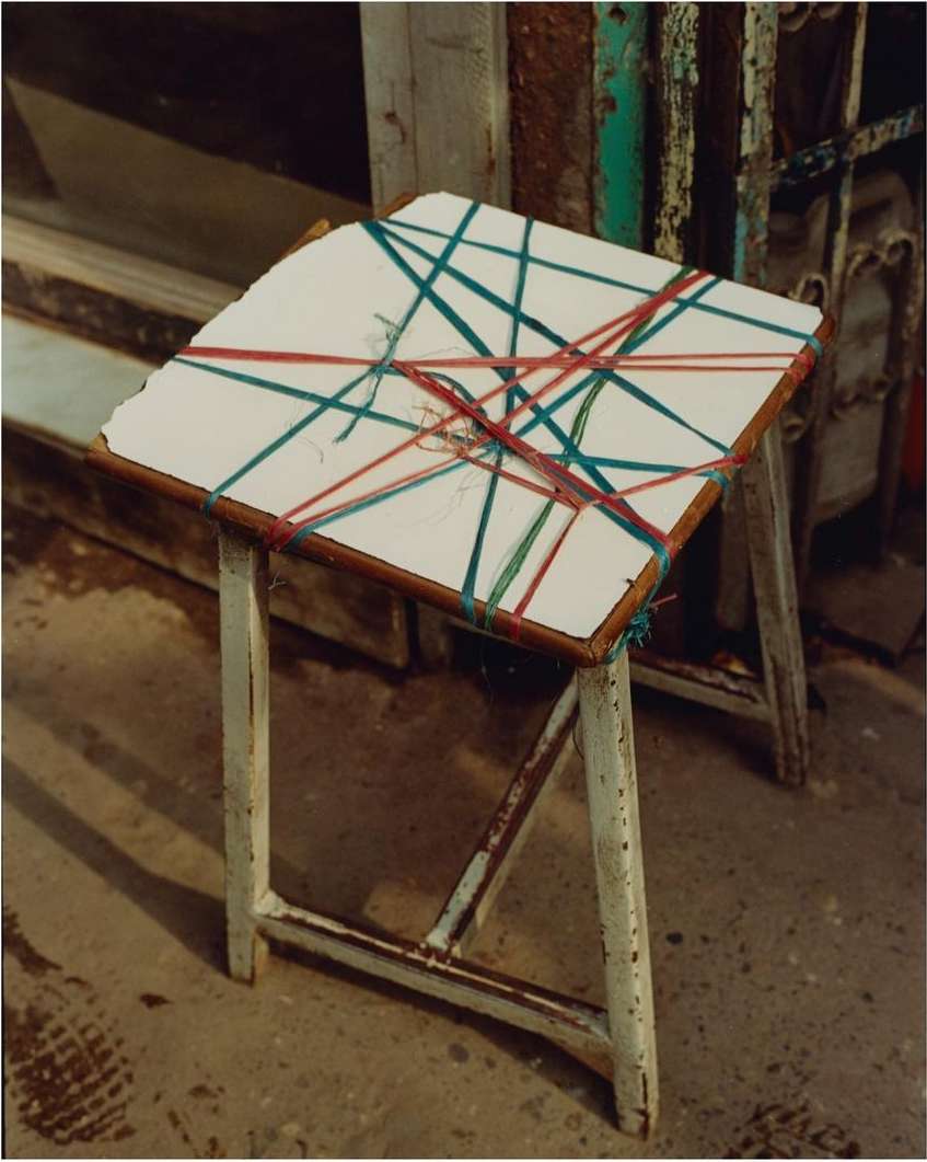 Rustic stool with white top crisscrossed by blue and red string sits outdoors on weathered concrete, a weathered building behind it.