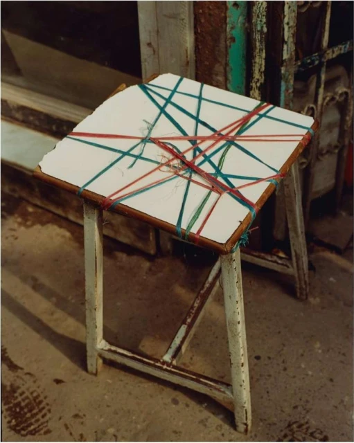 Rustic stool with white top crisscrossed by blue and red string sits outdoors on weathered concrete, a weathered building behind it.