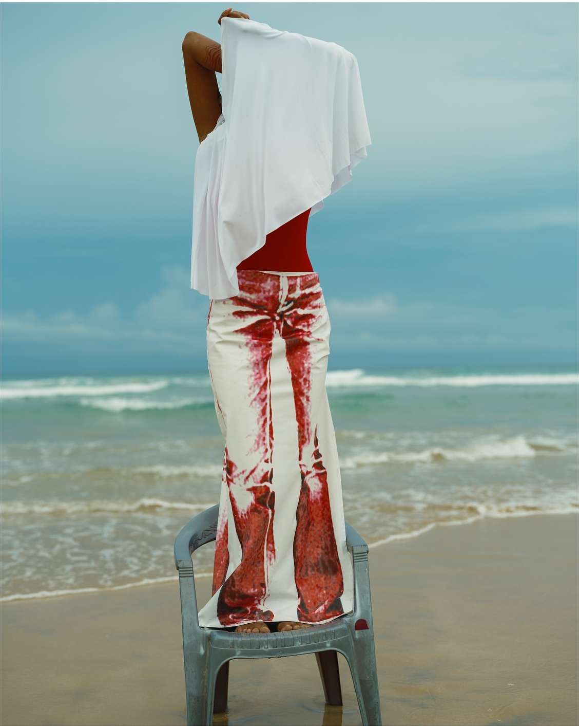 A woman stands on a beach in a printed skirt, holding a white cloth, with ocean waves and a cloudy sky creating a serene backdrop.