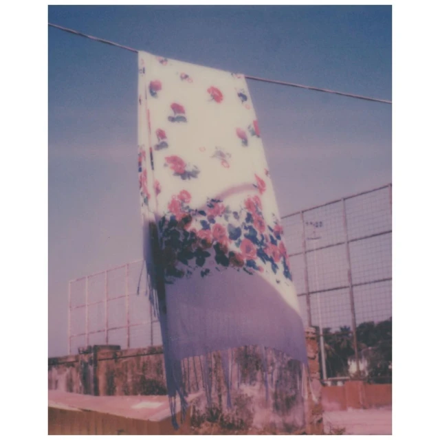 Floral shawl draped over a line on a sunny day with a building and vegetation in the background, creating a peaceful outdoor scene.