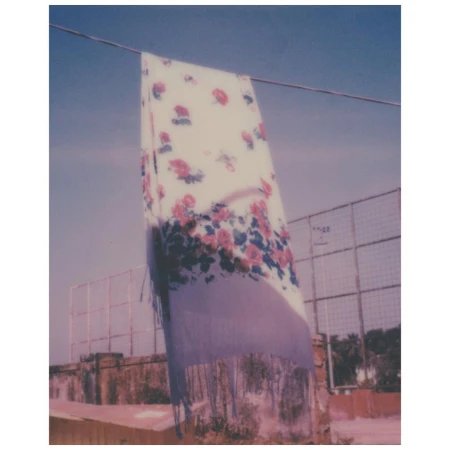 Floral shawl draped over a line on a sunny day with a building and vegetation in the background, creating a peaceful outdoor scene.