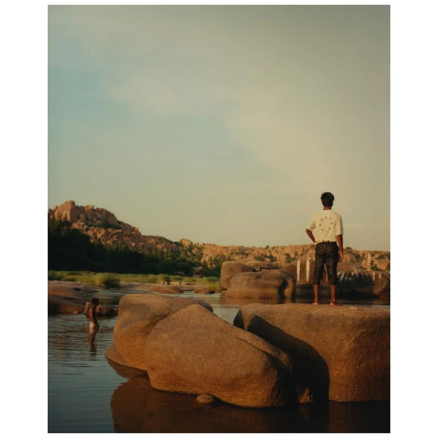Man standing on rocks in a river with other people wading, surrounded by rock formations under a blue sky, landscape orientation.