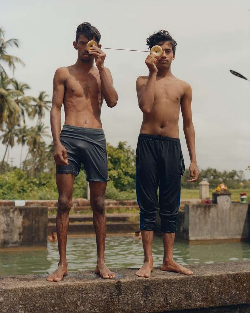 Two shirtless young men playfully pose by a pool, holding donut-shaped objects to their eyes, with a serene outdoor backdrop.