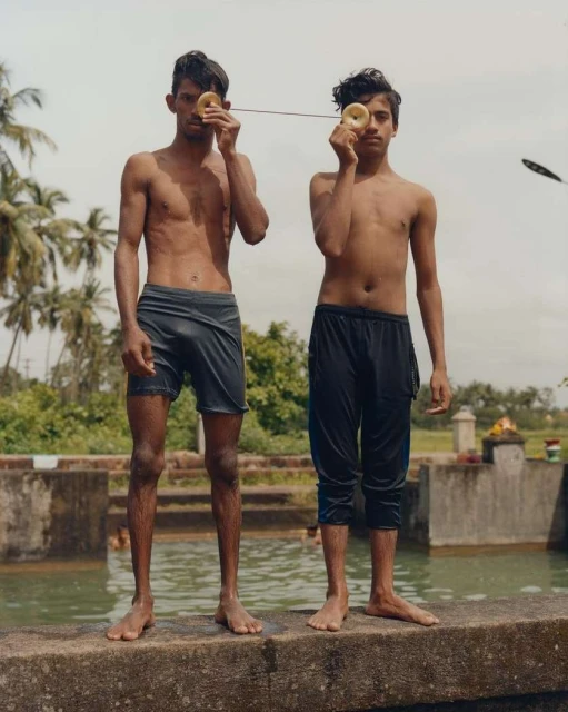Two shirtless young men playfully pose by a pool, holding donut-shaped objects to their eyes, with a serene outdoor backdrop.