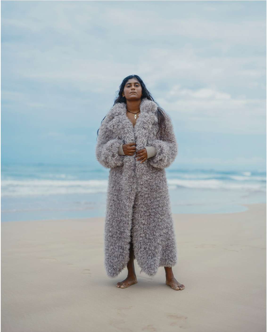 Woman in a fur coat stands barefoot on a sandy beach. Waves crash in the background under an overcast sky.