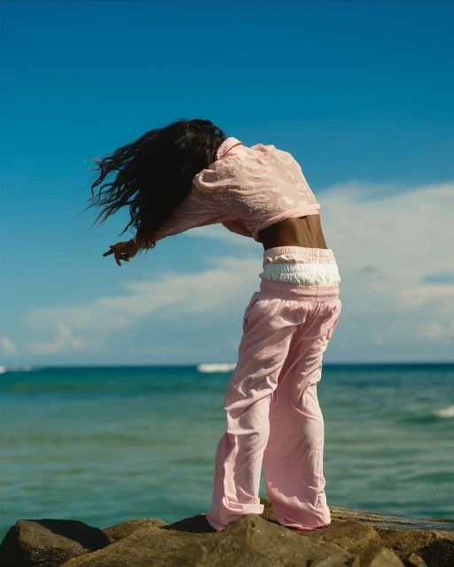 Adult woman in pink outfit bending over backwards outdoors on a rocky surface near ocean, blue sky in the background.