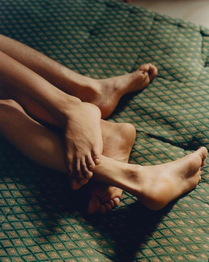 Close-up of the lower legs and feet of two people intertwined on a green patterned textile in an indoor setting with soft, diffused light.