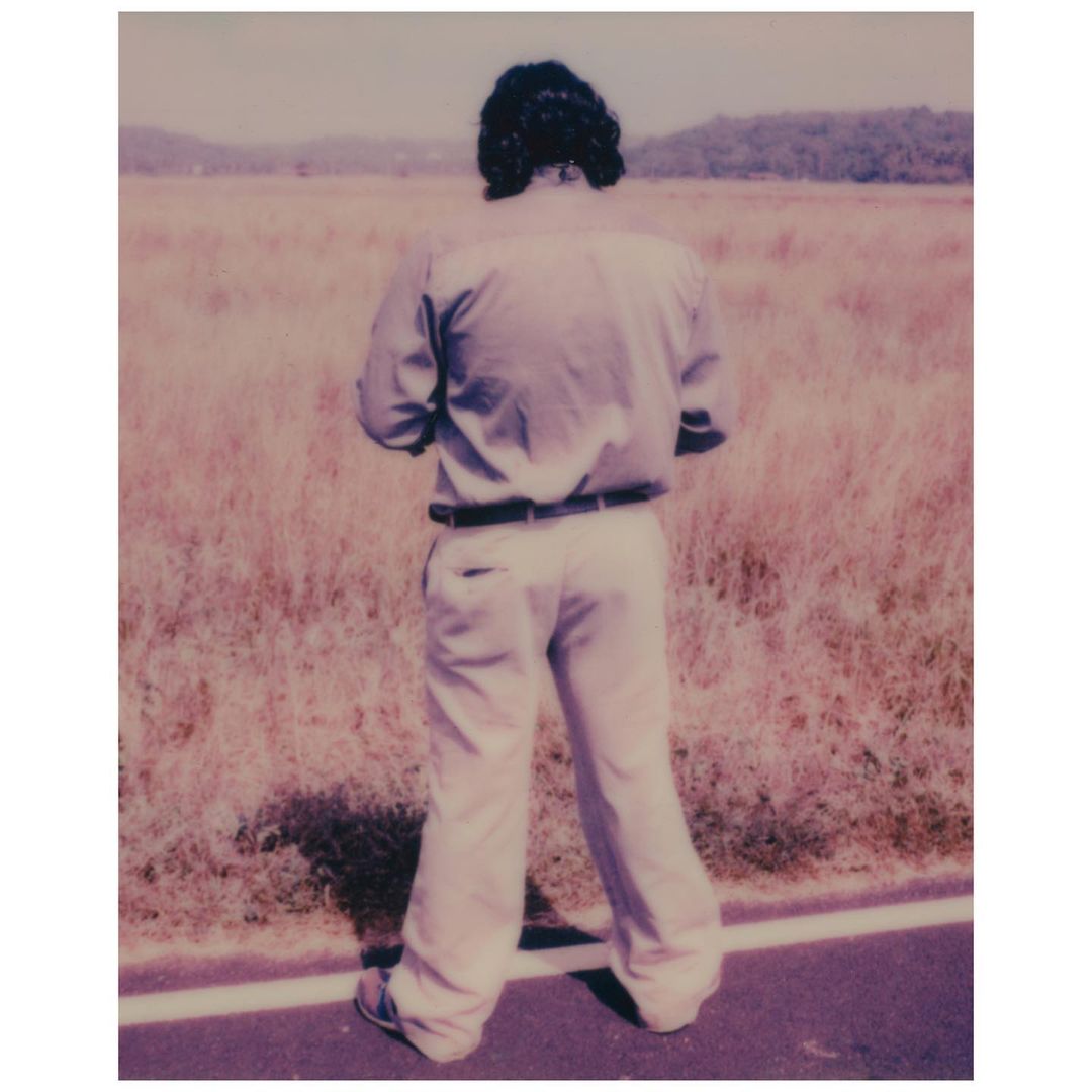 Back view of an adult in casual wear, standing on a road next to a field with distant trees beneath a clear sky on a sunny day.