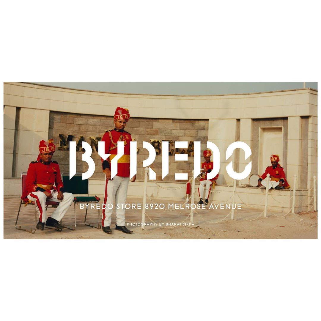 Five musicians in matching red uniforms pose outside a neutral-toned building promoting Byredo at 8920 Melrose Avenue, photographed by Bharat Sikka.