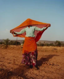 Person holding up an orange veil covering their face, wearing a light green top and patterned skirt in a dry field.
