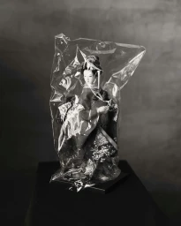 Traditional Japanese geisha doll encased in crinkled clear plastic wrapping resting on a dark surface in front of a textured gray wall.
