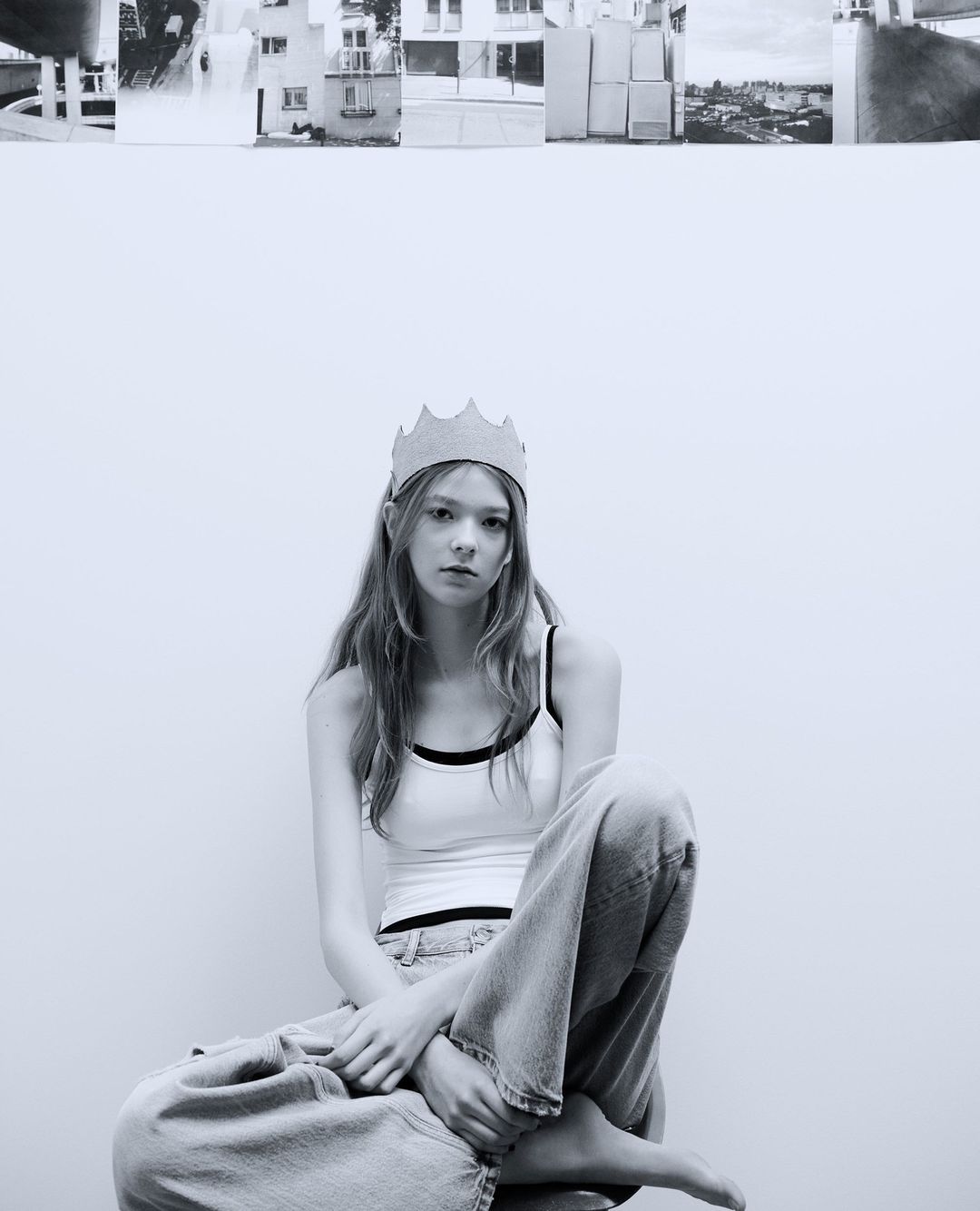 Black and white portrait of a young woman with crown. She sits on stool in front of a white wall, with architectural collage above.