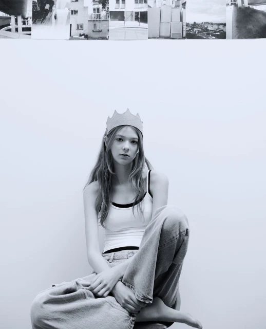 Black and white portrait of a young woman with crown. She sits on stool in front of a white wall, with architectural collage above.